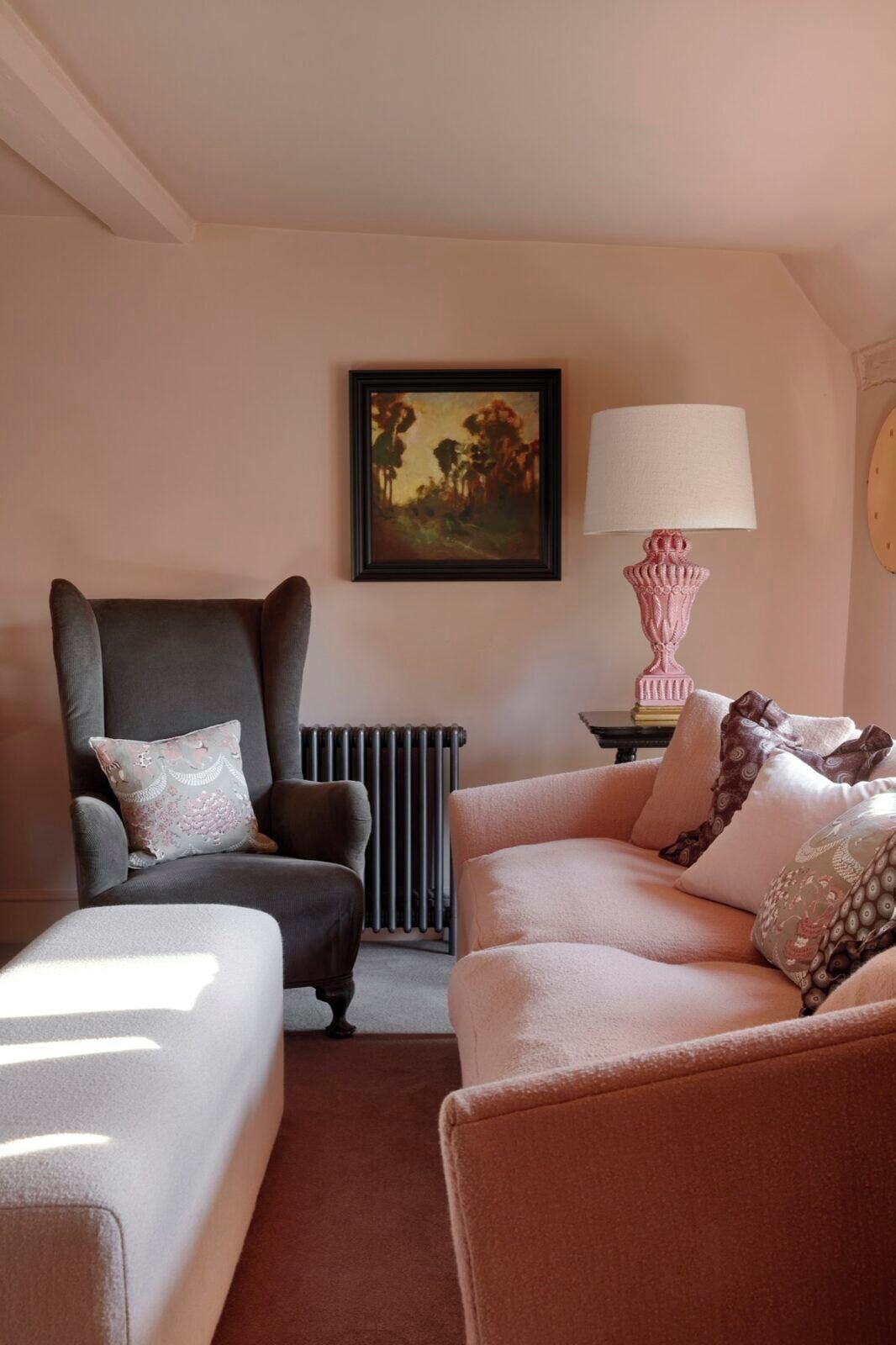 A cozy living room with a grey armchair, a pink sofa, and a white sofa. There's a radiator, a framed painting, and a pink lamp on a side table.
