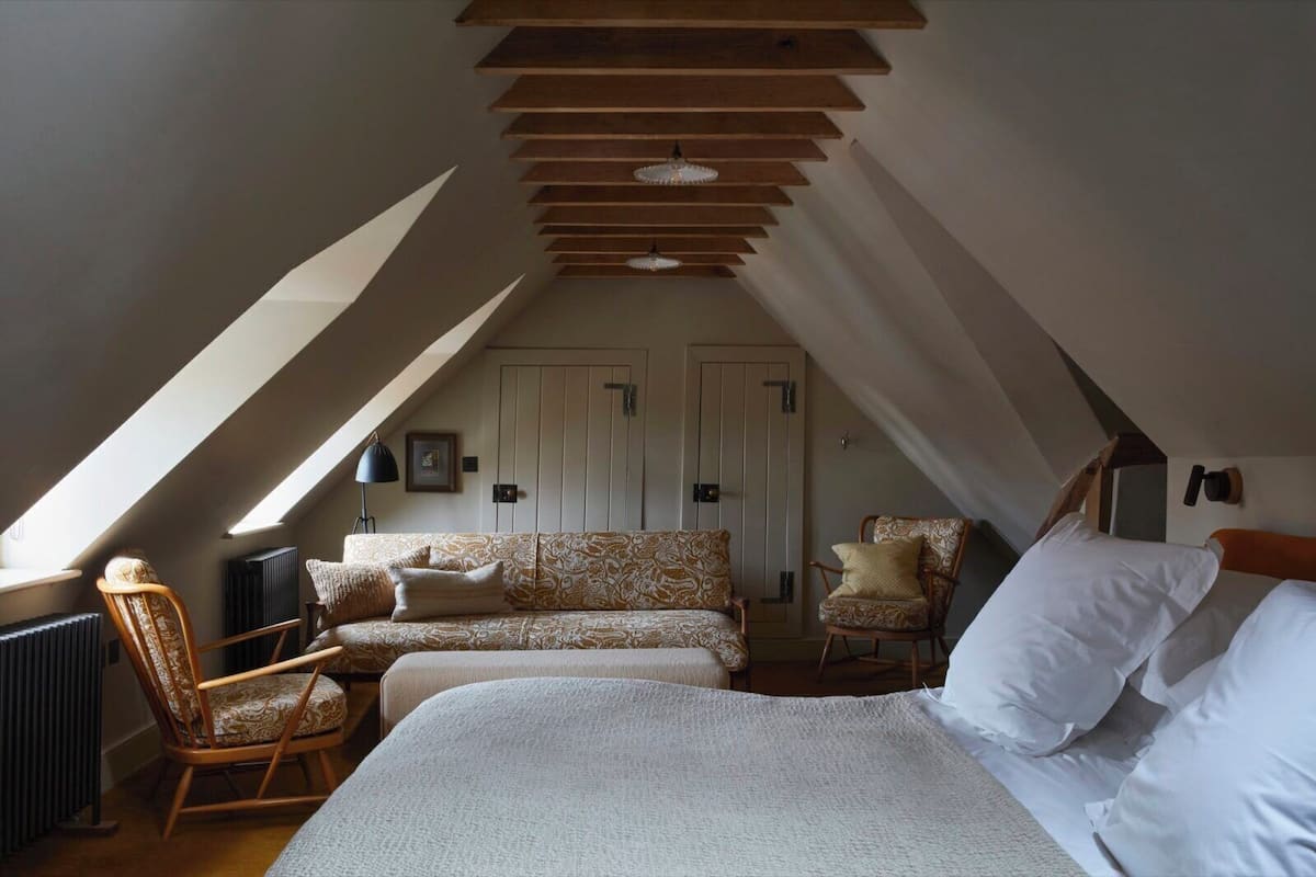 Un dormitorio acogedor en el ático, con techo inclinado, una cama grande, dos sillones, un sofá y un radiador.