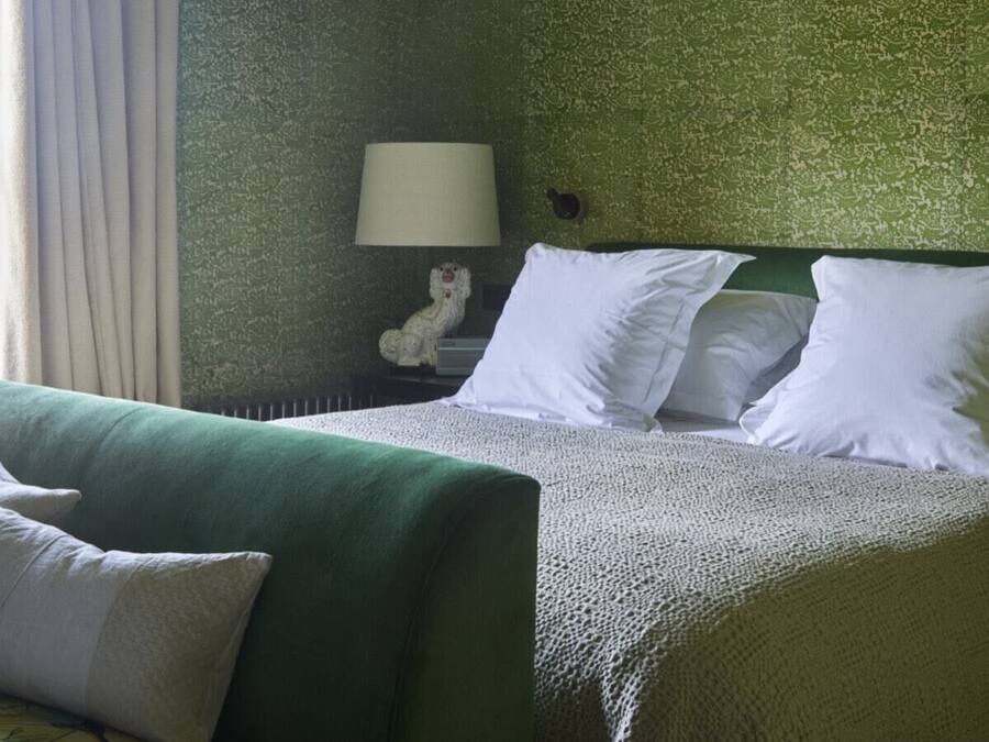 Habitación de hotel con pared verde, cama con ropa blanca, mesita de noche con lámpara y ventana con cortinas.