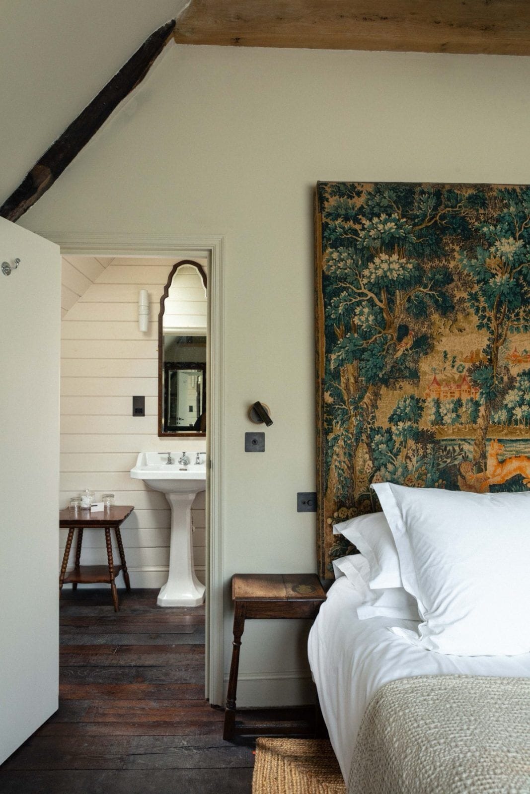 A bedroom with a bed, a nightstand, a wooden bench, a white sink, and a mirror in a hallway.