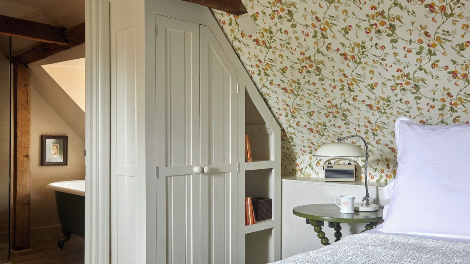 Un dormitorio con techo inclinado, un armario blanco, una mesa verde con lámpara y un estampado floral en el papel tapiz.