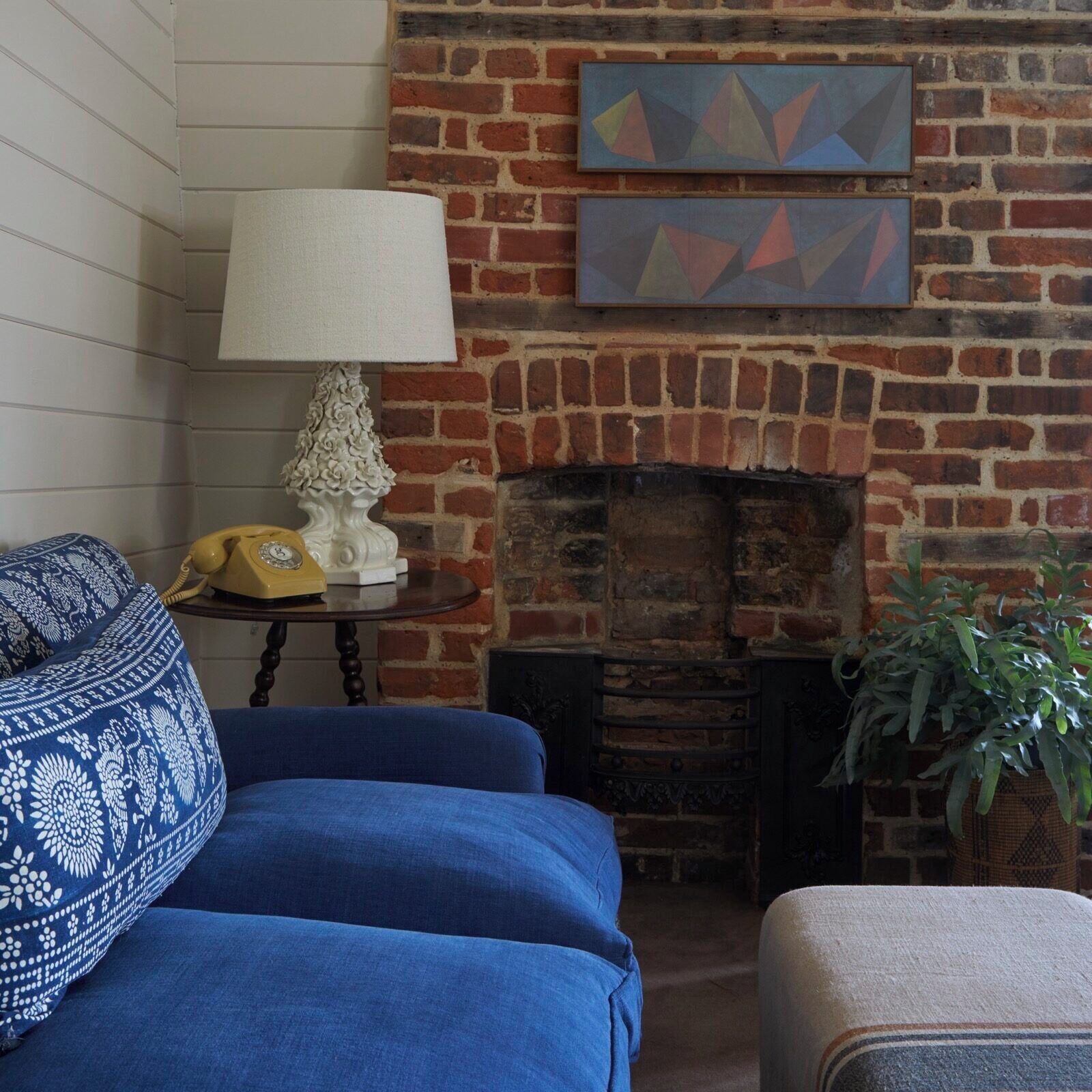 A cozy living room with a brick fireplace, a blue sofa, a patterned pillow, a white lamp, and a telephone on a small table.