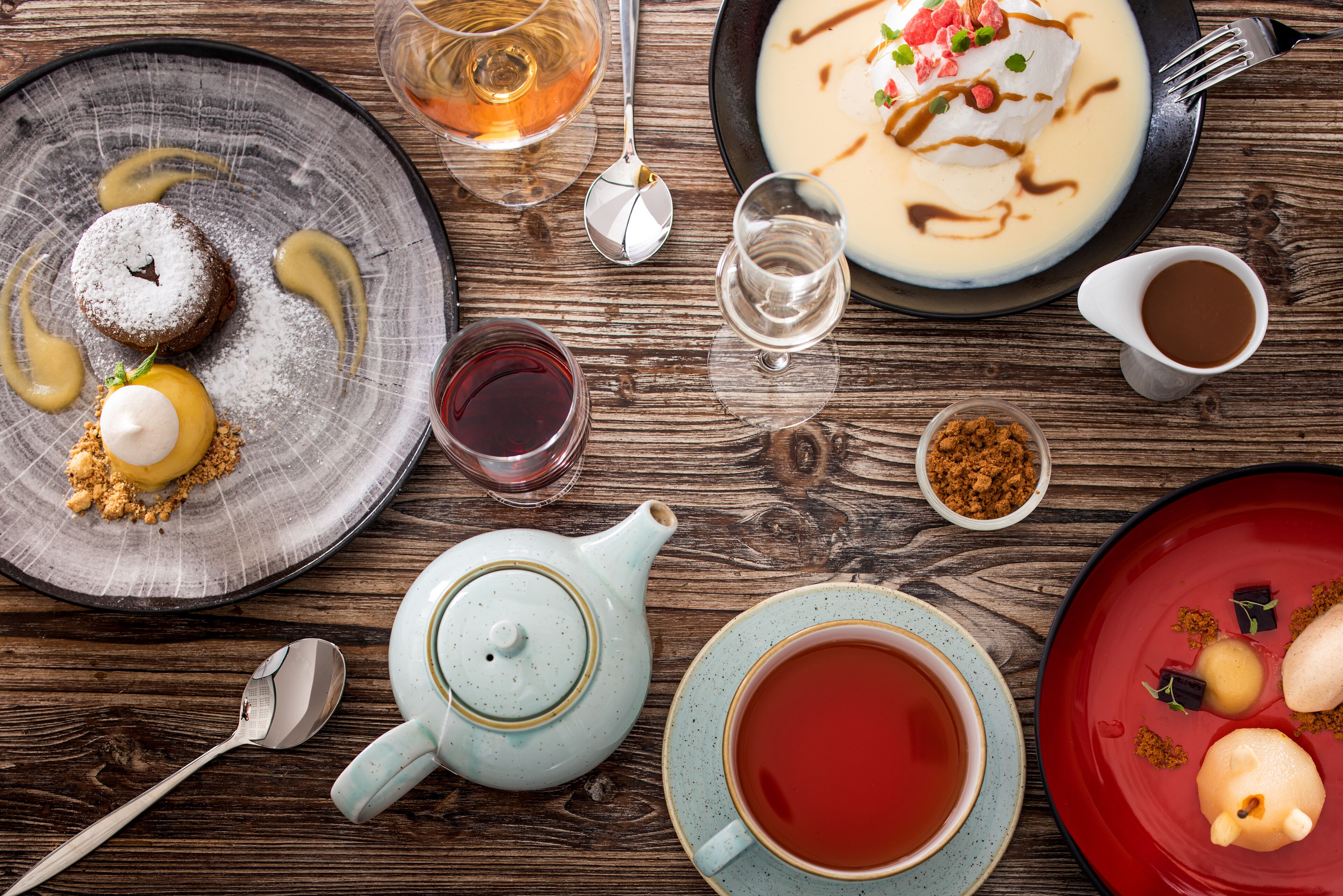 Una mesa de madera con diversos platos, incluyendo un postre con azúcar glas, un tazón de sopa con una cucharada de crema, y una tetera con una taza de té.