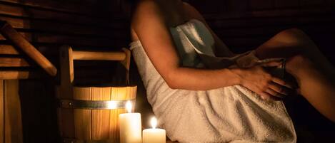 A person sitting on a wooden bench in a dimly lit room with two lit candles and a wooden bucket.