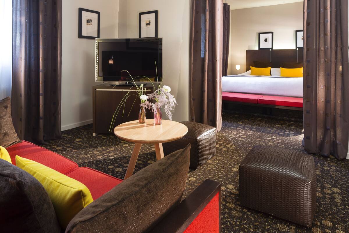 A hotel room with a sofa, ottoman, a small round table with a vase, a television, and a bed with yellow pillows.