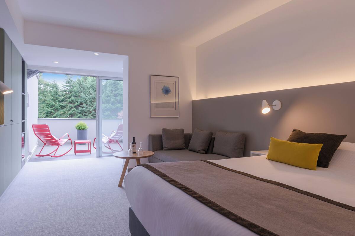 A modern hotel room with a large bed, a sofa, a small table with a plant, and a red chair.