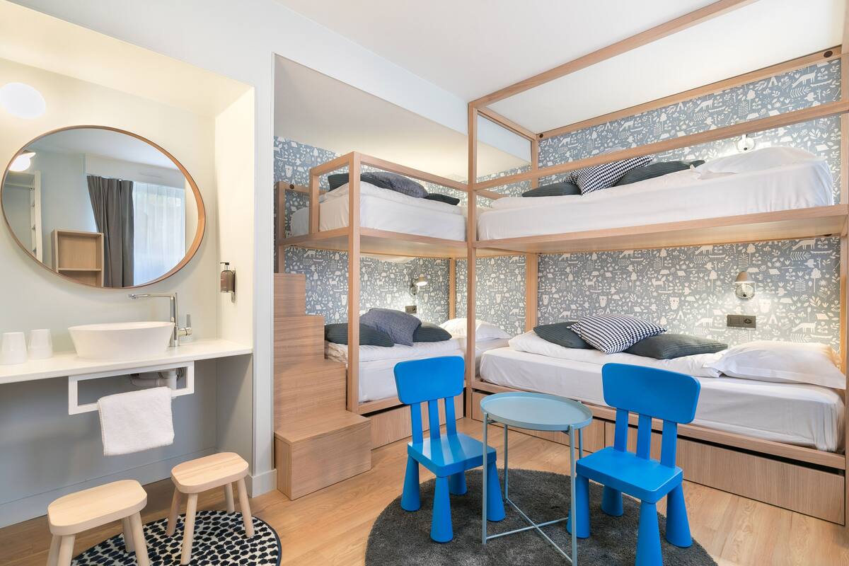 A compact hotel room with bunk beds, a round mirror, a small round table, and two blue chairs.