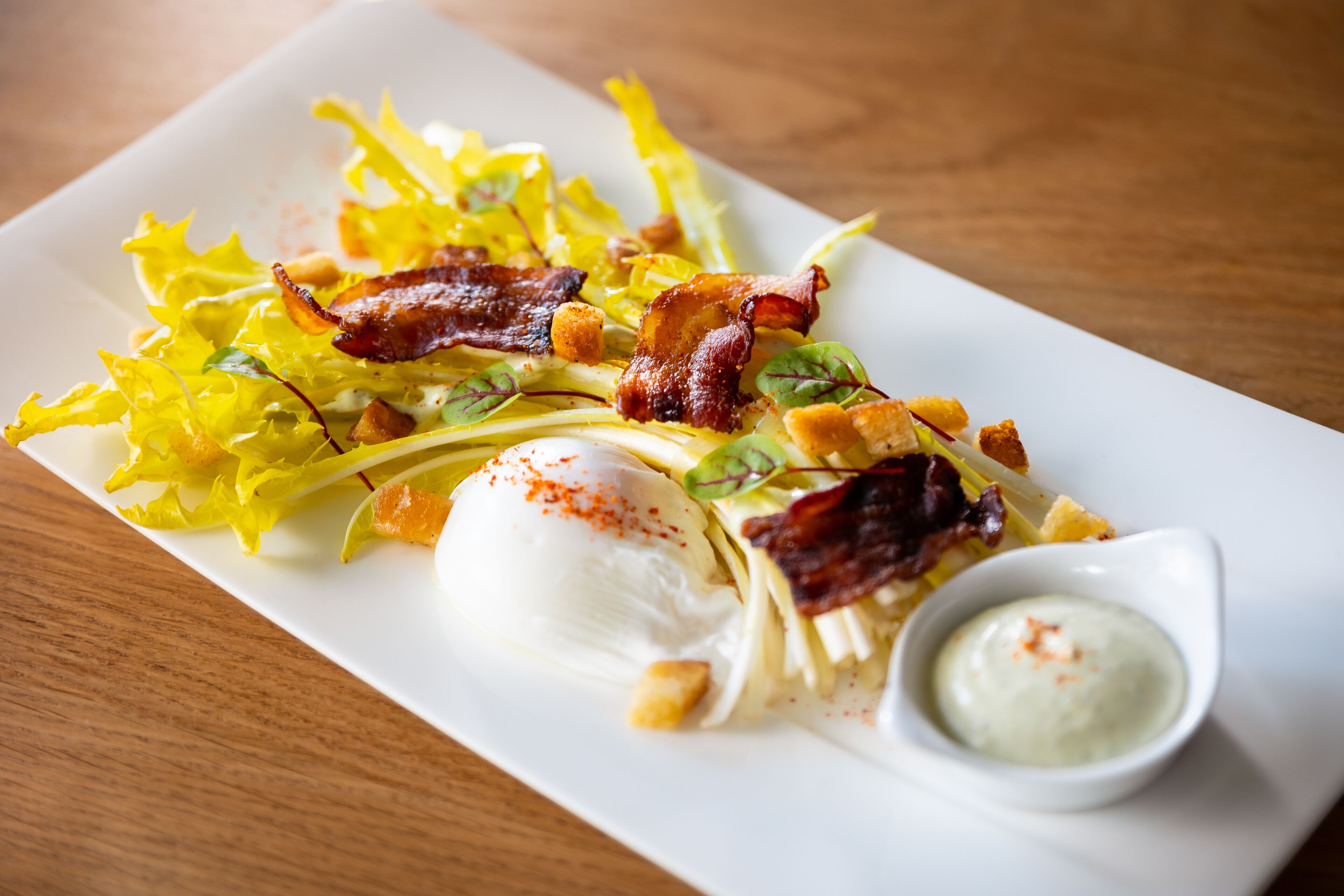 Un plato rectangular blanco con ensalada, un huevo escalfado, tocino crujiente, crotones y un recipiente pequeño con salsa.