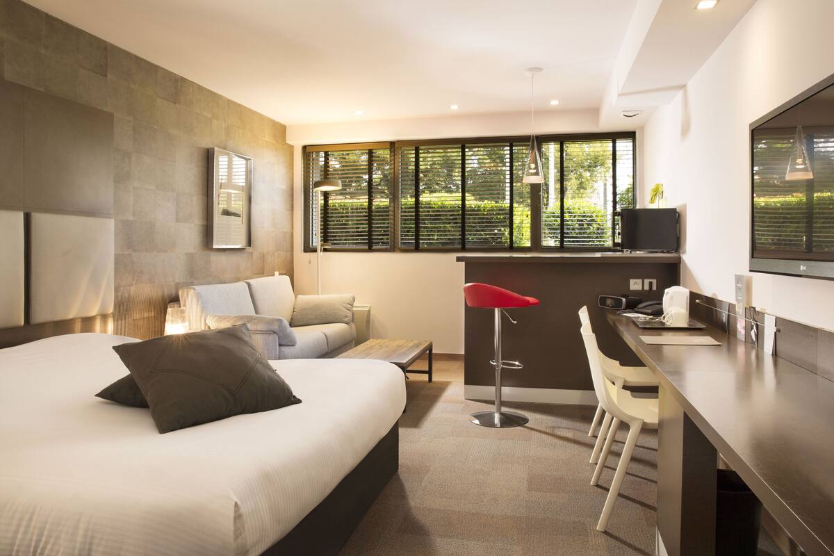 A modern hotel room with a large bed, a desk, and a view of greenery through the window.