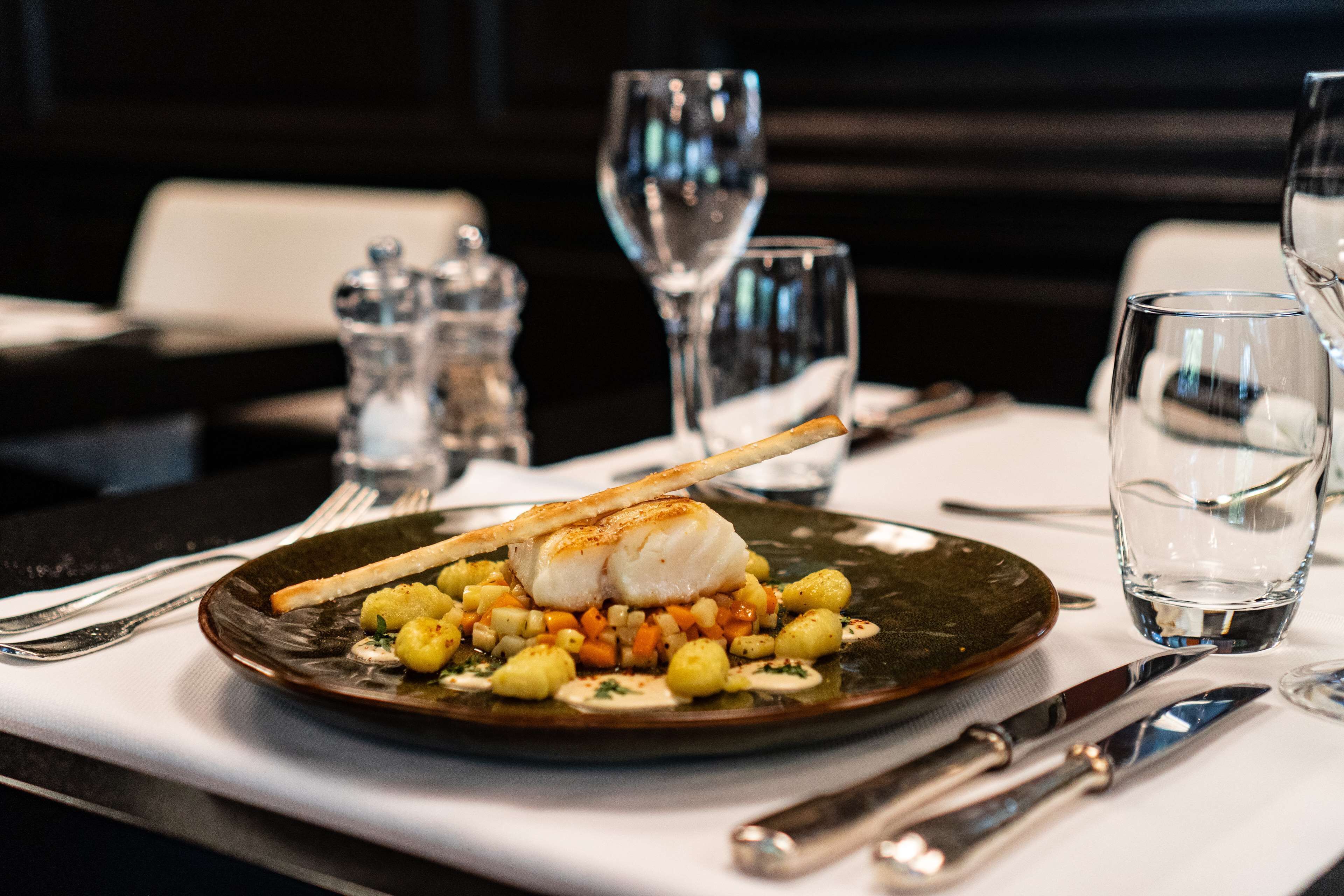 A plate of food with a fish, vegetables, and a piece of bread on a dark plate. There are glasses and cutlery on the table.