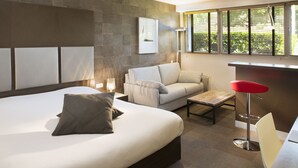 Quarto de hotel moderno com uma cama grande, um sofá, uma pequena mesa e um banquinho vermelho.