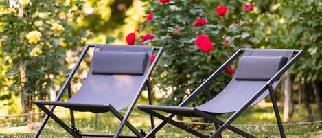 Two lounge chairs in a grassy outdoor area with a backdrop of green foliage and red flowers.