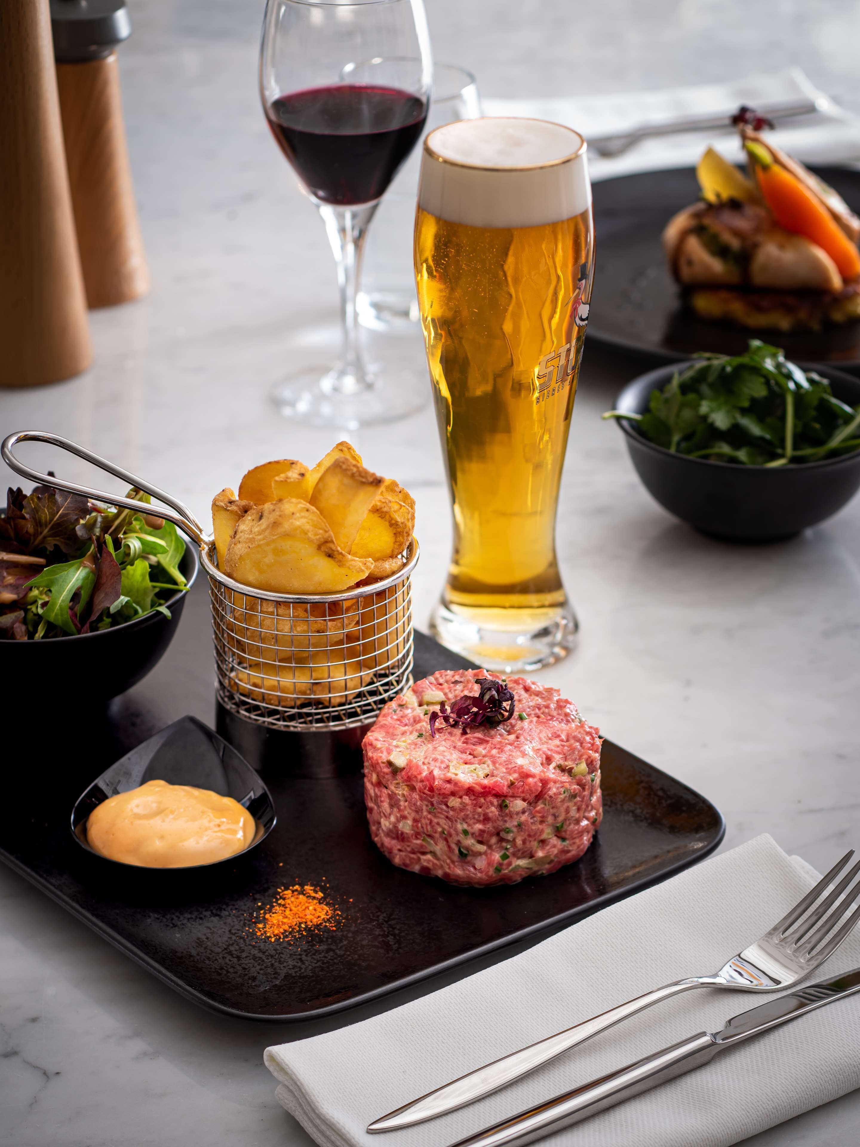 A dining table with a variety of food items including a steak tartare, a glass of beer, a bowl of salad, and a plate of chips.
