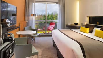 A hotel room with a bed, a desk, a chair, a TV, and a balcony with a view of trees.