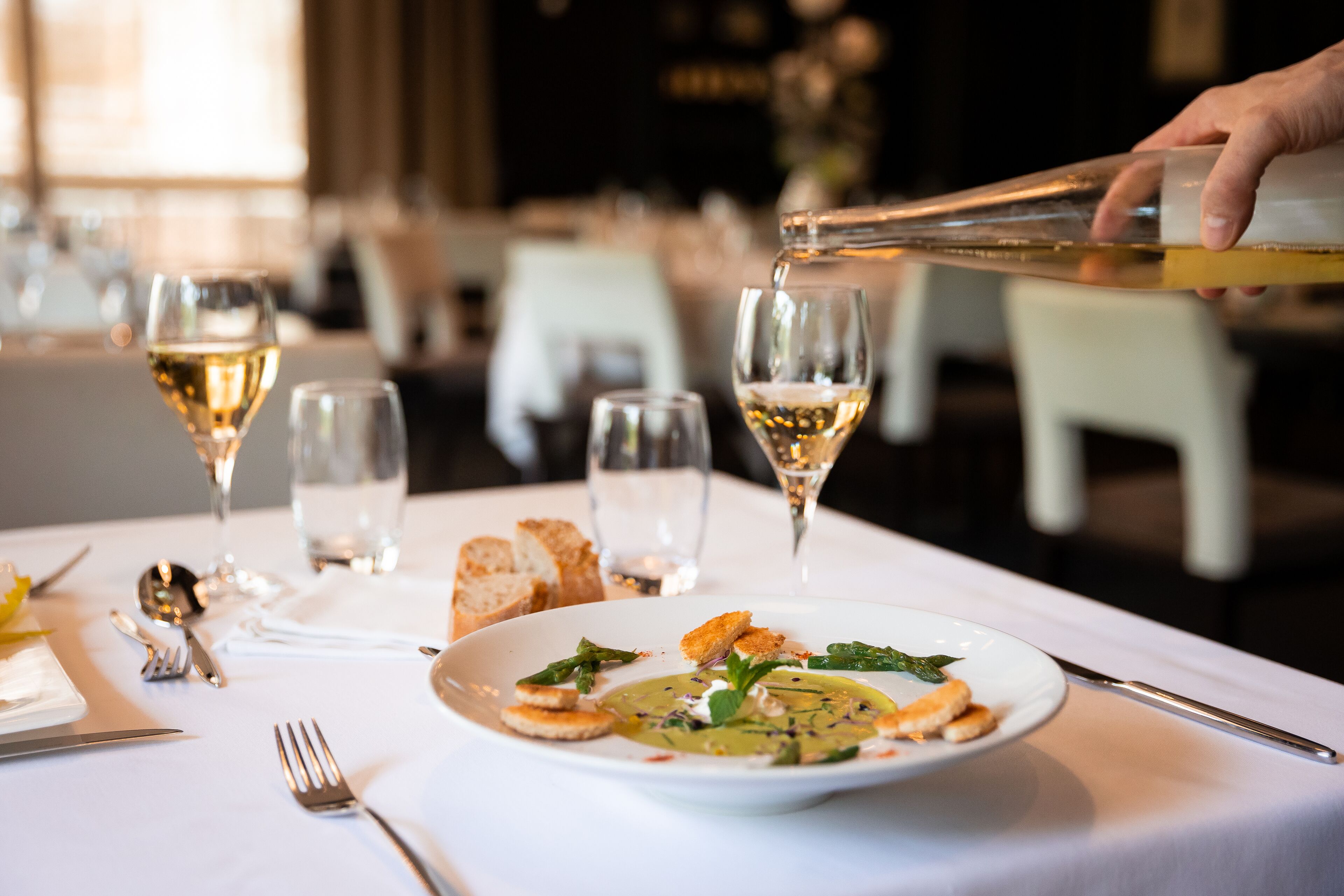 Une table à manger avec une assiette de nourriture, des verres à vin et une bouteille de vin dont on verse le contenu.