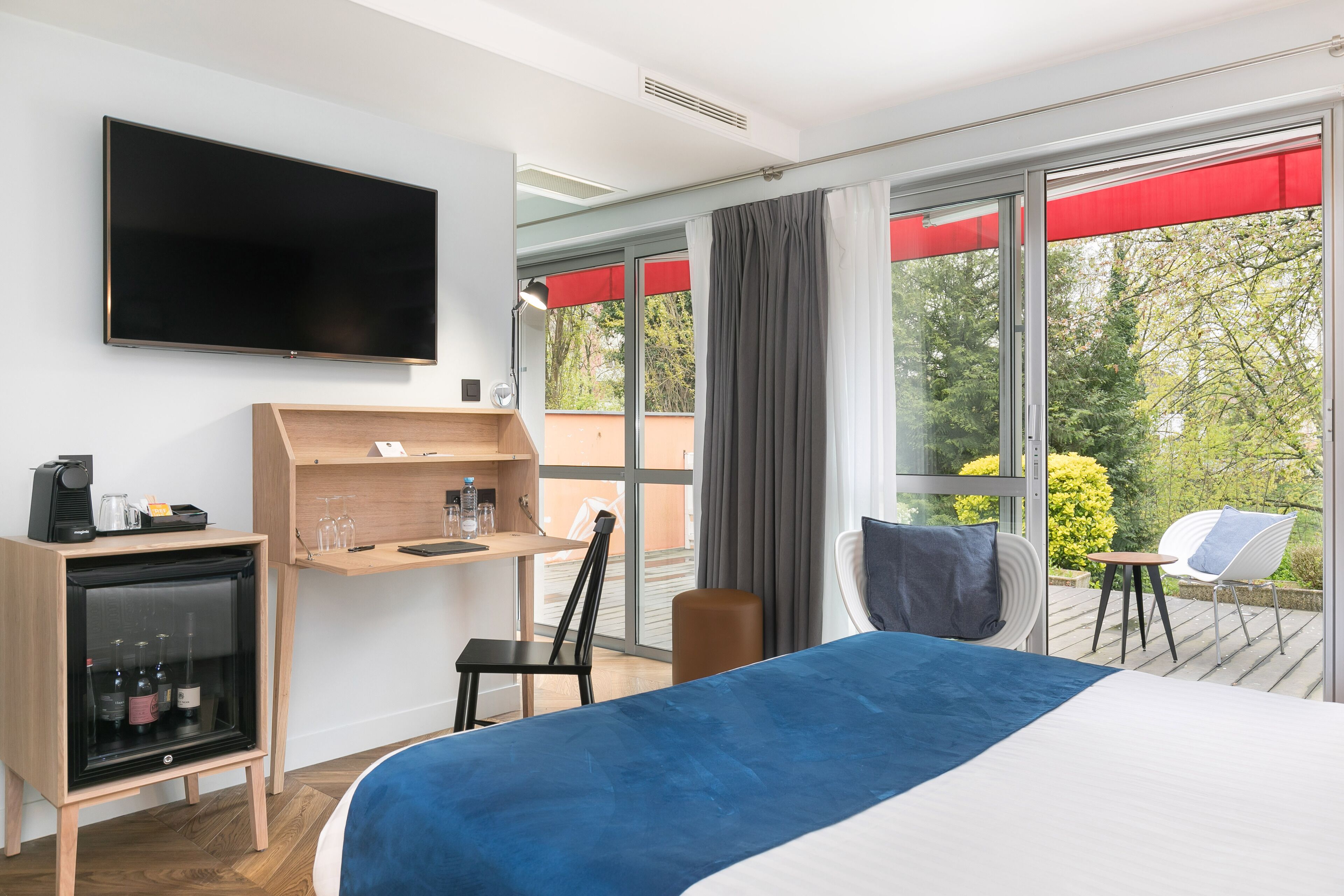 A modern hotel room with a large bed, a desk with a computer, a TV, and a balcony with a chair and table.