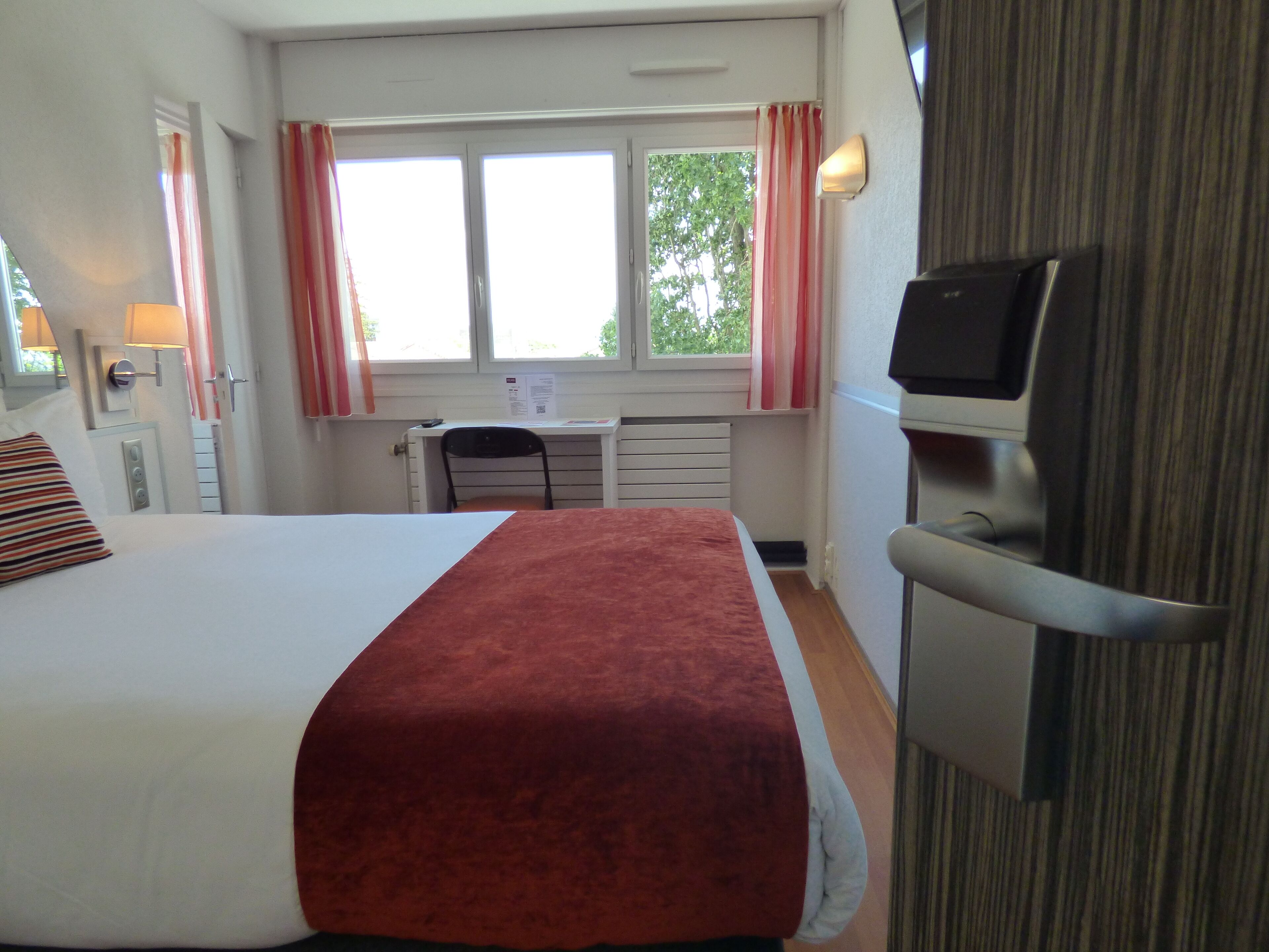 Habitación de hotel con cama, escritorio, silla, televisor y ventana con cortinas.