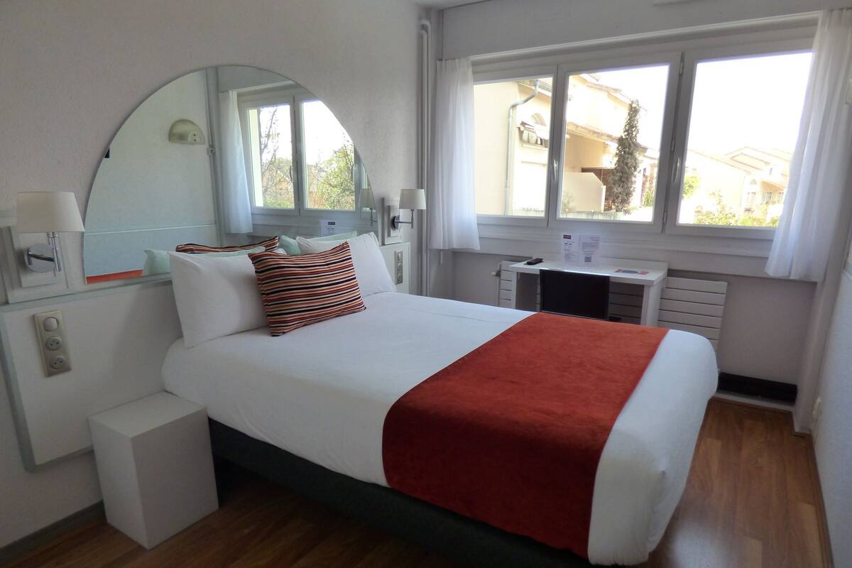 A hotel room with a bed, a desk, a chair, a mirror, and a window.
