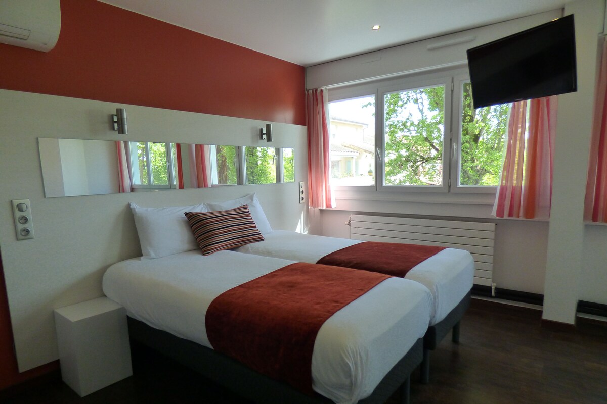 A double bed with a red throw blanket, a bedside table, a flat-screen TV, and a window with curtains.