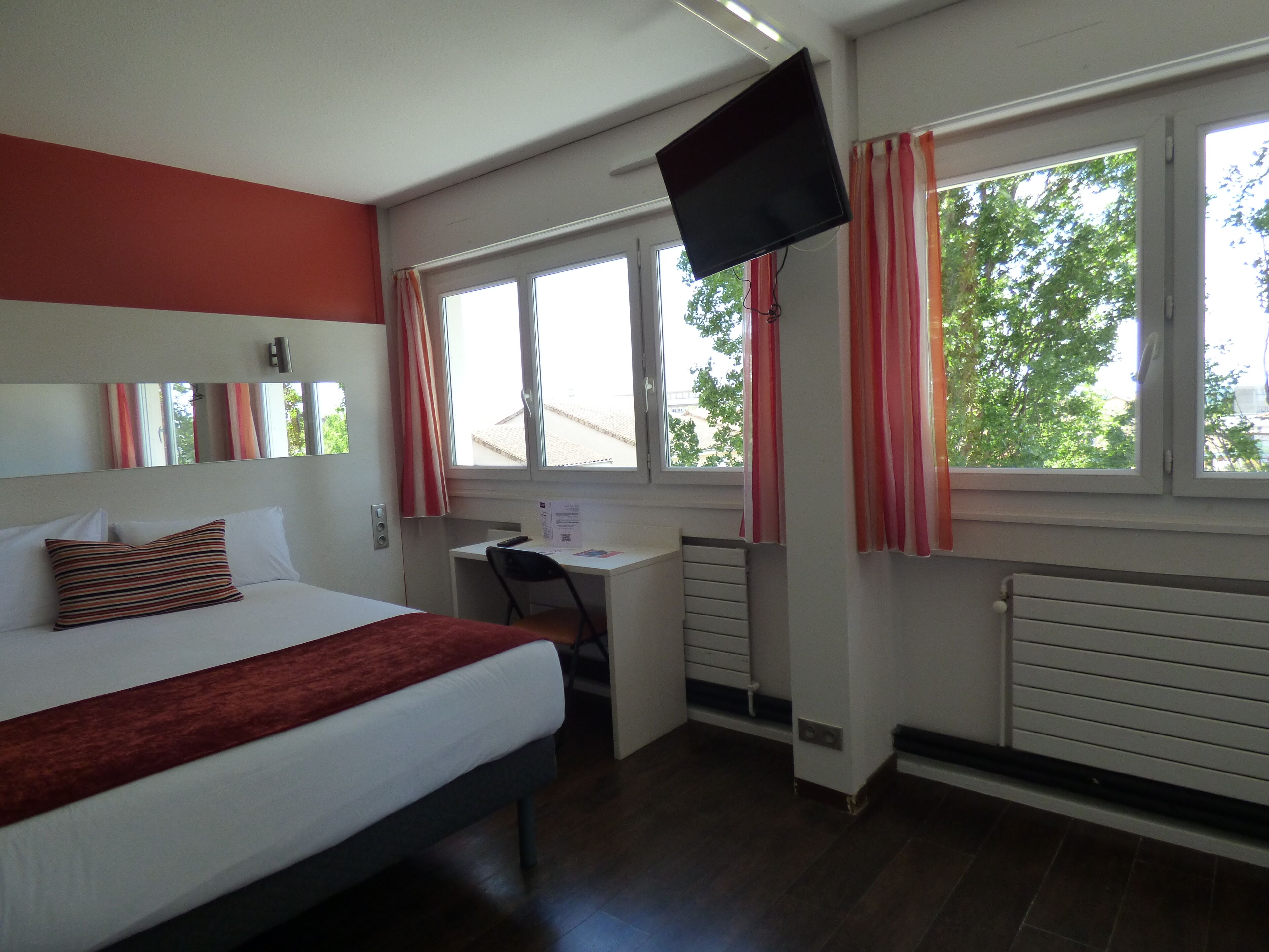 Une chambre d’hôtel équipée d’un lit, d’un bureau, d’une chaise, d’une télévision et de fenêtres avec des rideaux.