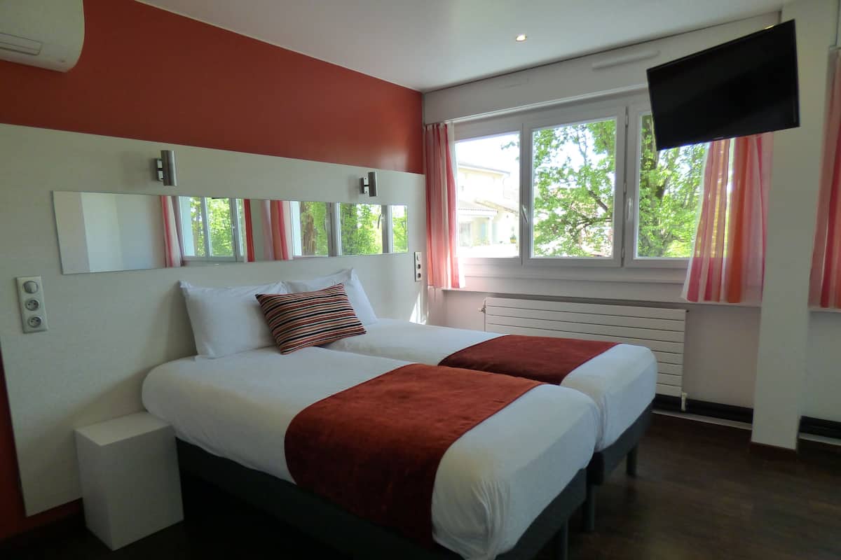 A double bed with a red throw blanket, a bedside table, a flat-screen TV, and a window with curtains.