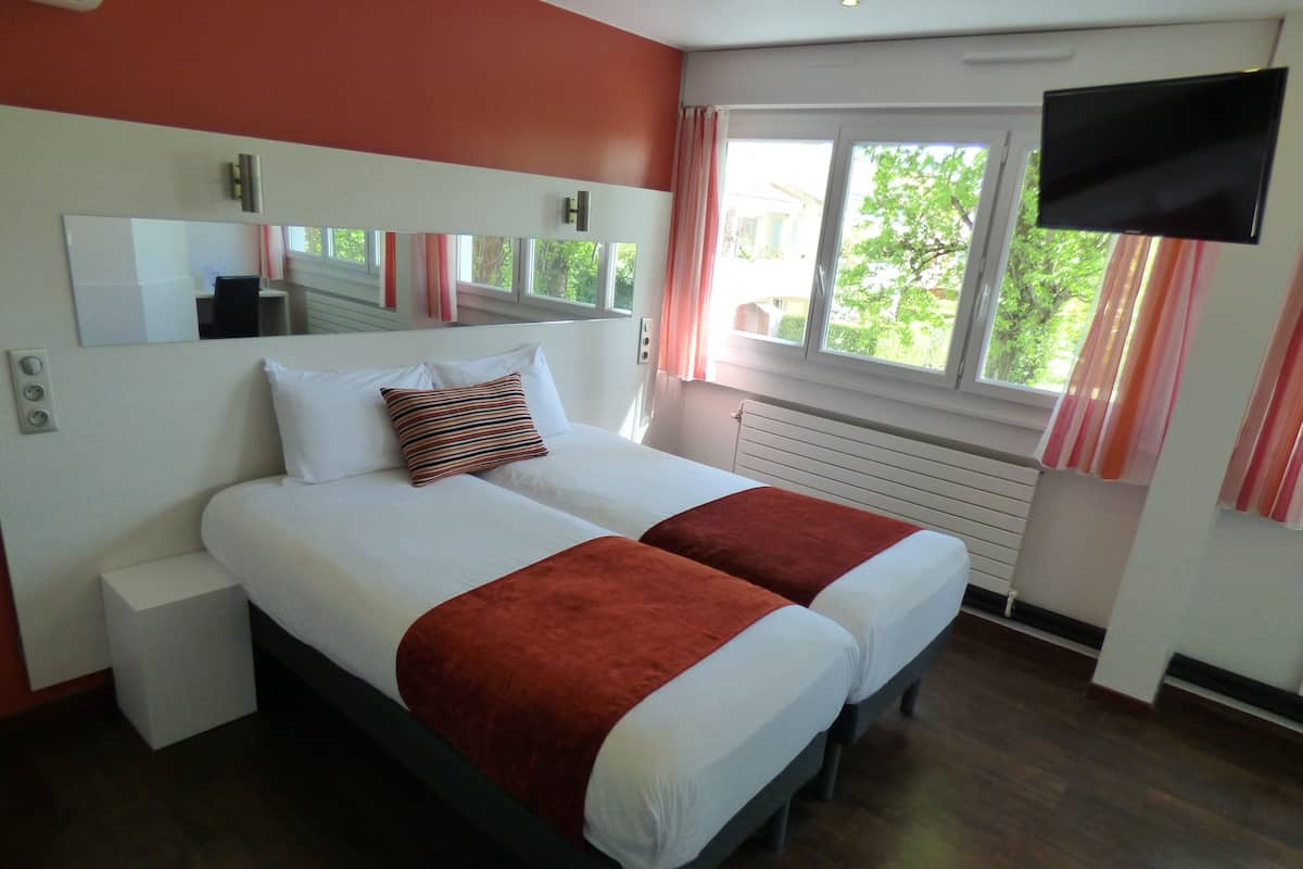 A double bed with white and red bedding, a bedside table, a large mirror, a flat-screen TV, and a window with curtains.
