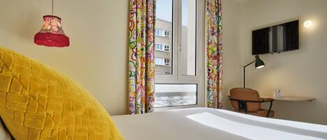 A hotel room with a bed, a desk, a chair, a TV mounted on the wall, and colorful curtains.