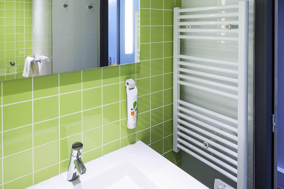 Une salle de bain avec un lavabo blanc, des murs recouverts de carreaux verts, un miroir et un porte-serviettes blanc.