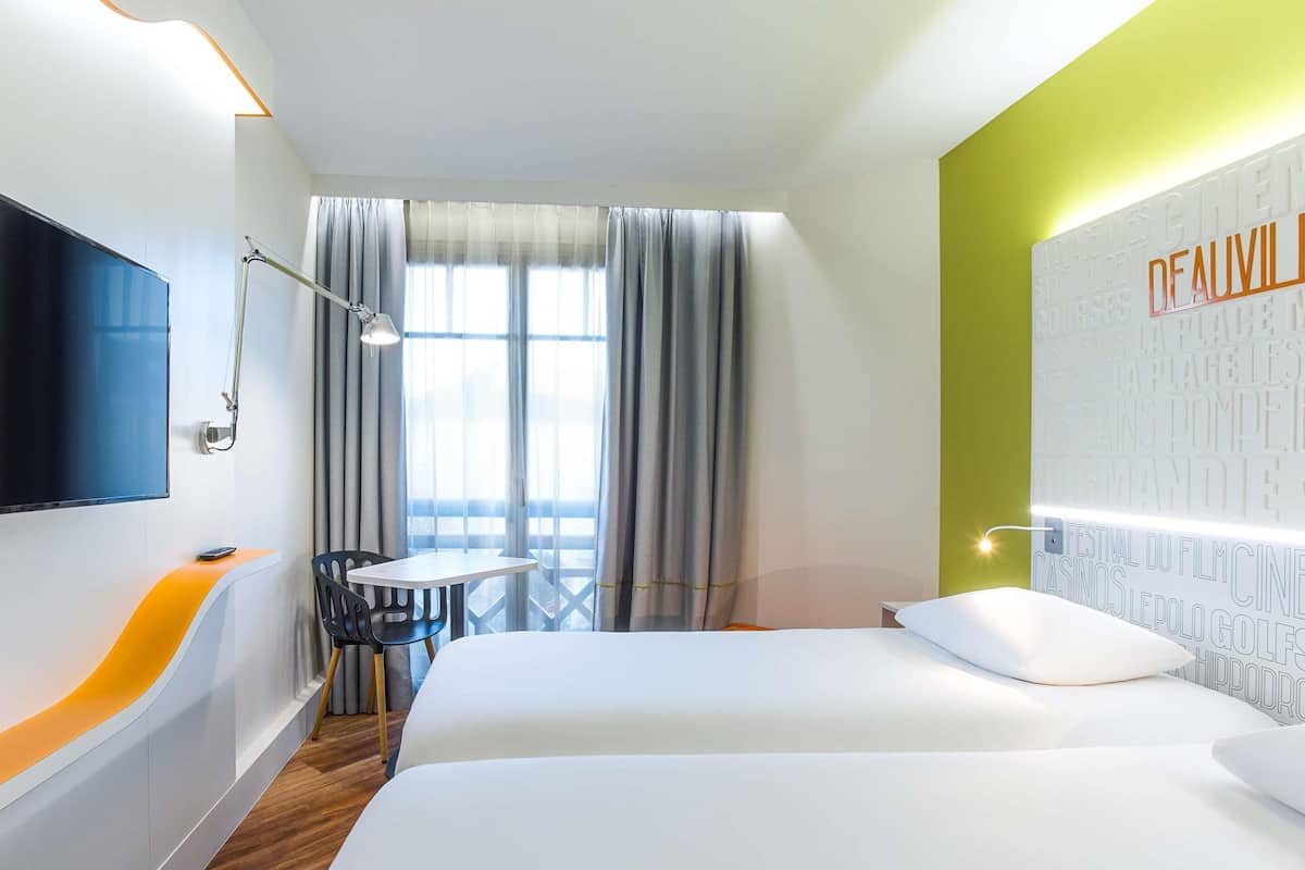A hotel room with a bed, a desk with a chair, a TV, and a window with curtains.