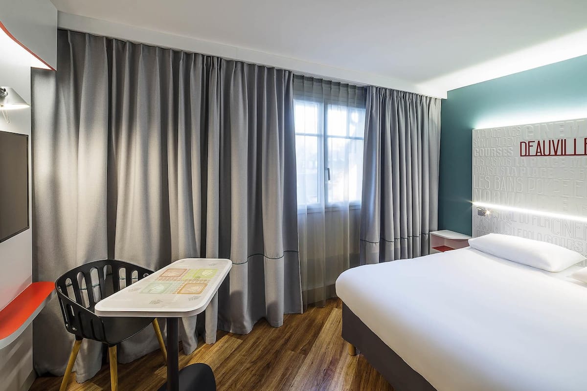 Une chambre d’hôtel avec un lit, une petite table, une chaise, une fenêtre avec des rideaux et un mur orné de texte.