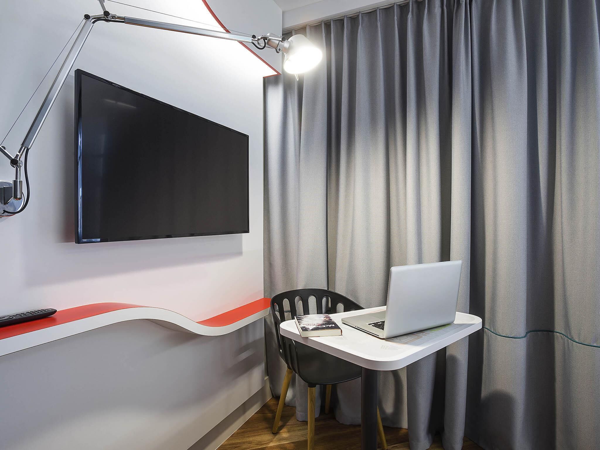 Une chambre moderne équipée d’un téléviseur à écran plat fixé au mur, d’un bureau avec un ordinateur portable, d’une chaise et d’une télécommande posée sur un comptoir rouge et blanc.