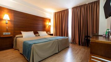 Ein Hotelzimmer mit massivem Holzkopfende, einem Bett mit beigefarbener Bettwäsche, zwei Nachttischlampen, einem Schreibtisch mit Flachbildfernseher und einem Fenster mit braunen Vorhängen.
