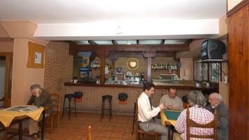 A bar with a brick wall, wooden beams, and a counter with various bottles and a television. There are several patrons seated at tables.