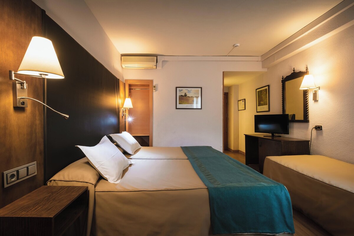 A hotel room with a bed, bedside tables, a TV, and wall-mounted lamps.