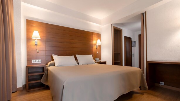 A hotel room with a large bed, wooden headboard, two bedside lamps, a nightstand, and a wooden wardrobe. - Hotel Corregidor (Segovia)