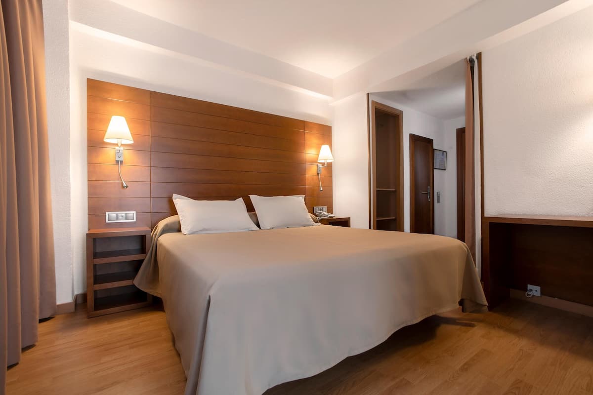 Habitación de hotel con una cama grande, cabecera de madera, dos lámparas de noche, una mesita de noche y un armario de madera.