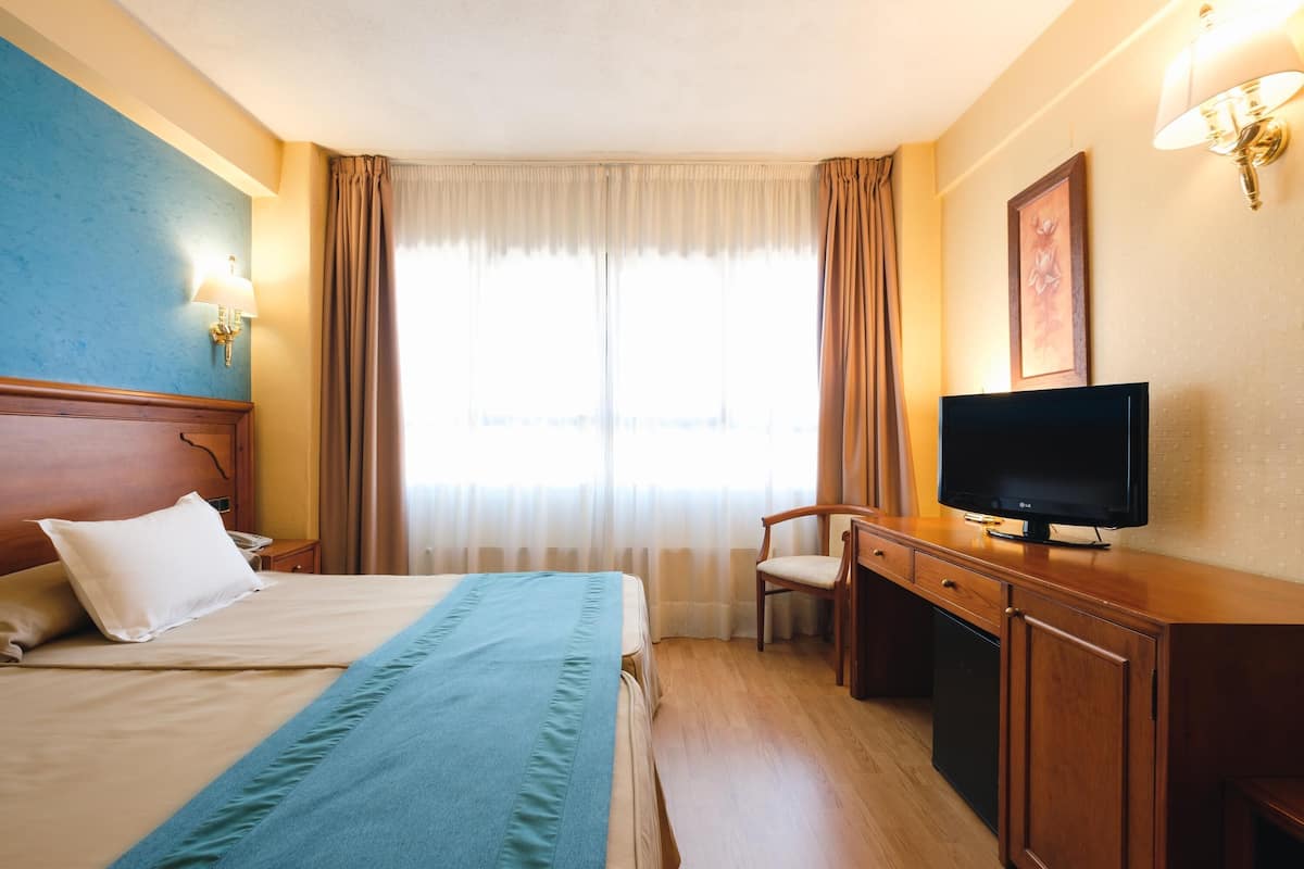 Una habitación de hotel con cama, escritorio, televisor y ventana con cortinas.