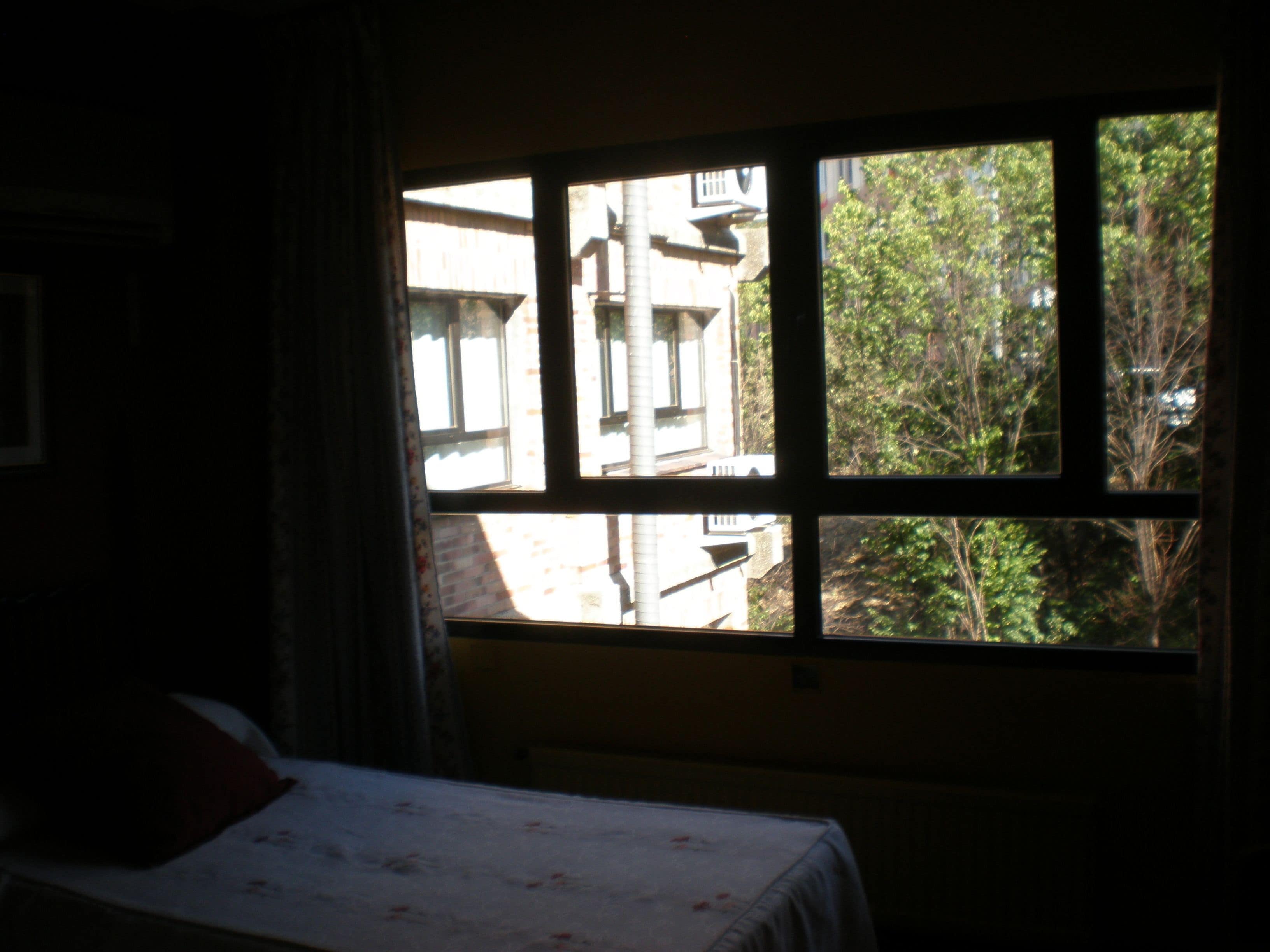 Quarto com cama, janela e vista para edifícios e árvores lá fora.