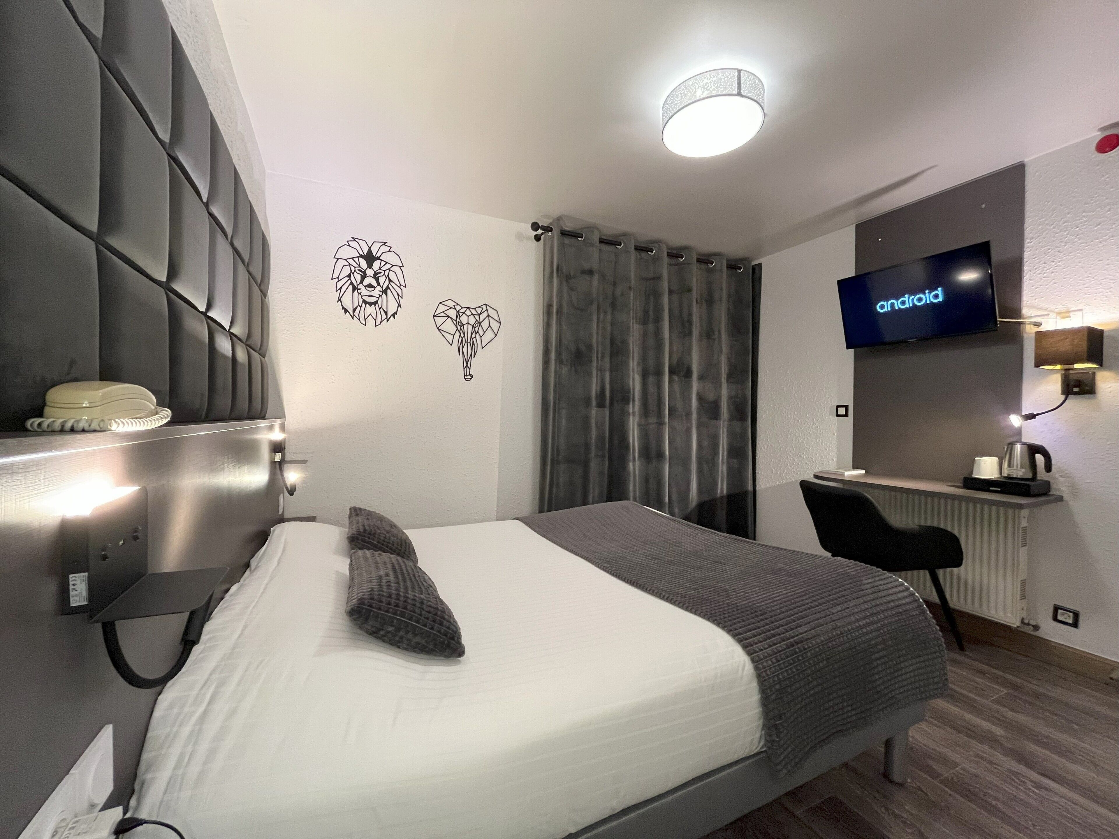 A modern hotel room with a bed, a desk, a television, and a wall-mounted light fixture.
