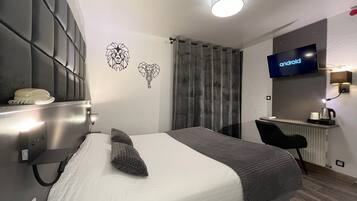 A modern hotel room with a bed, a desk, a television, and a wall-mounted light fixture.