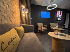 Un moderno vestíbulo de hotel con pared de madera, una pantalla de bienvenida azul, sillones estampados, una mesa redonda de madera y un sofá con un cojín 'Cognac'.