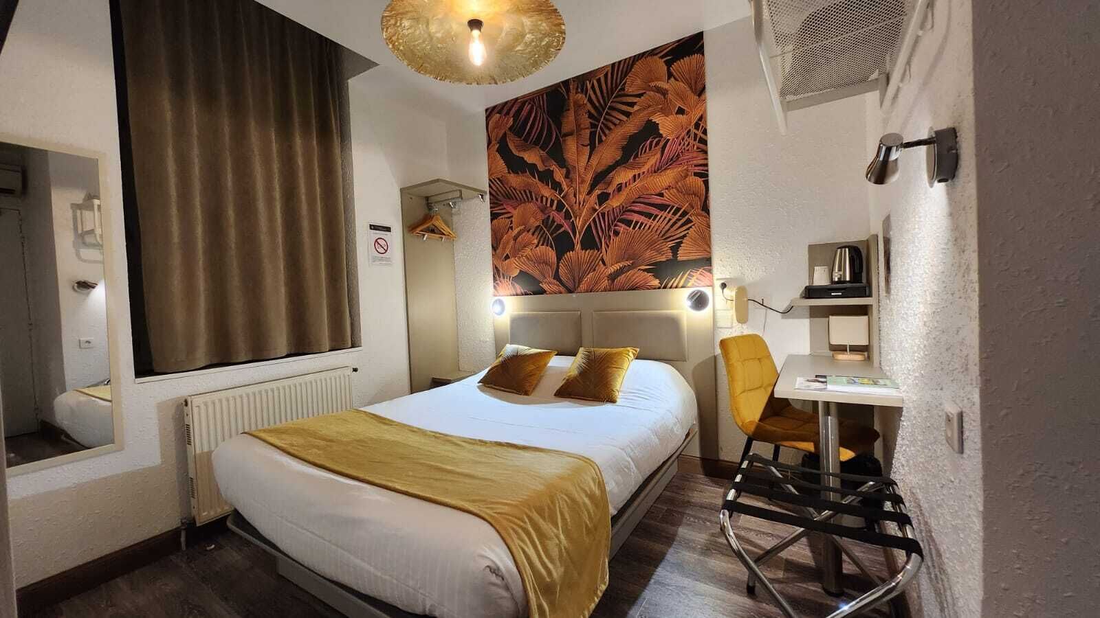 A hotel room with a bed, a desk, a chair, a radiator, a window with curtains, a wall-mounted light fixture, and a wall art piece.