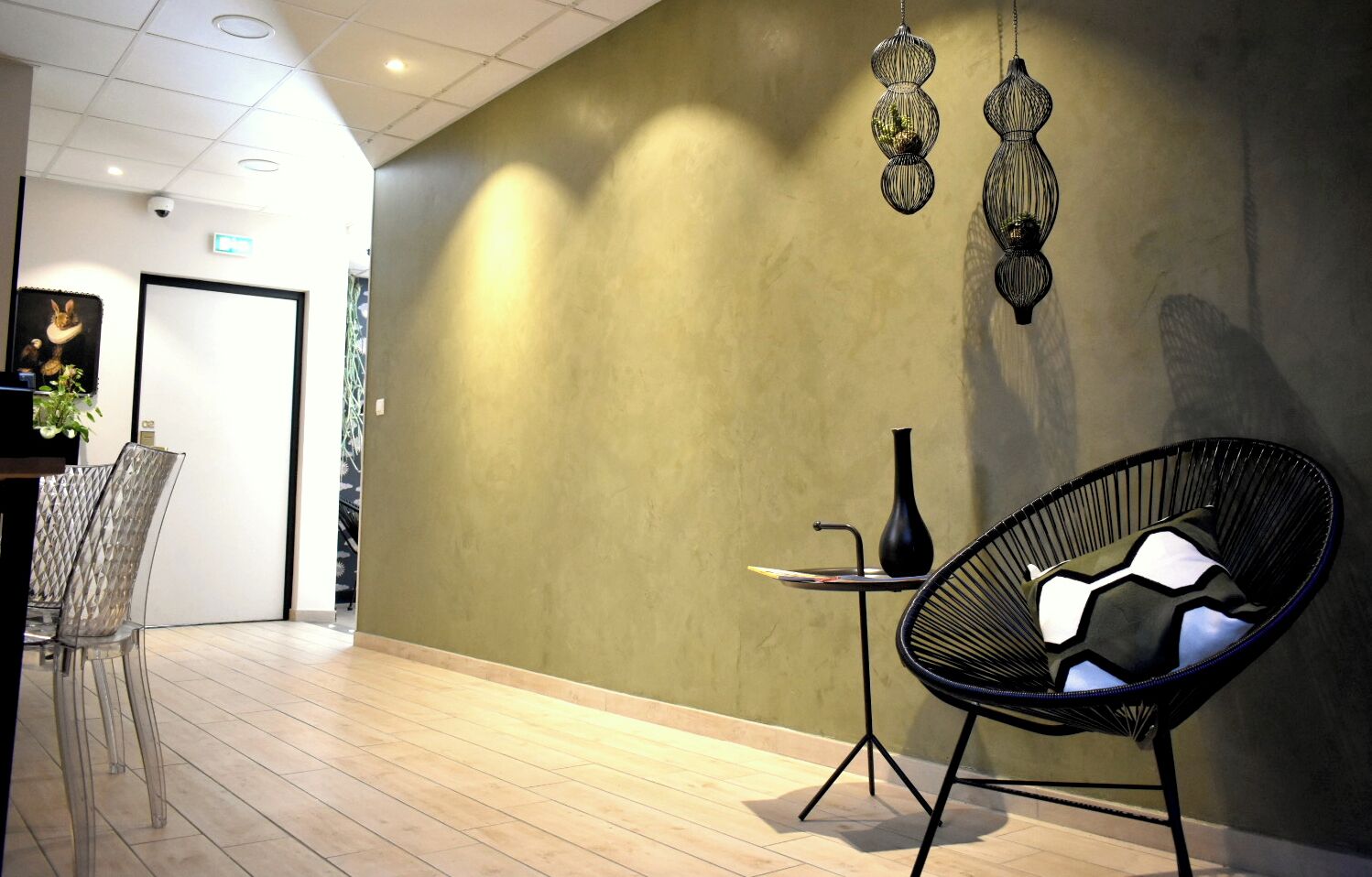 Ein modernes Interieur mit einer gewölbten Wand, einem schwarzen Stuhl aus Draht, einem kleinen Tisch mit einer Vase und einer Tür zu einem weiteren Raum.
