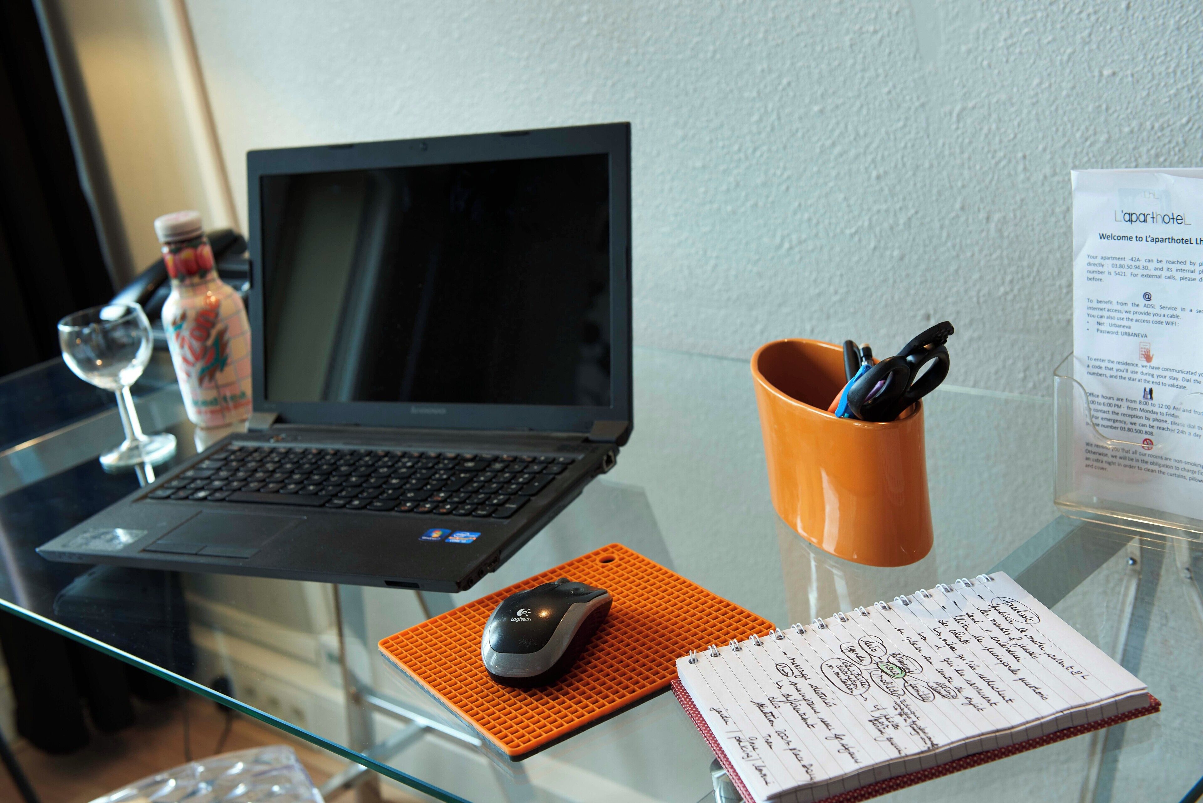 A laptop on a glass table, a notebook with handwritten notes, a mouse, a bottle, and a glass of water.