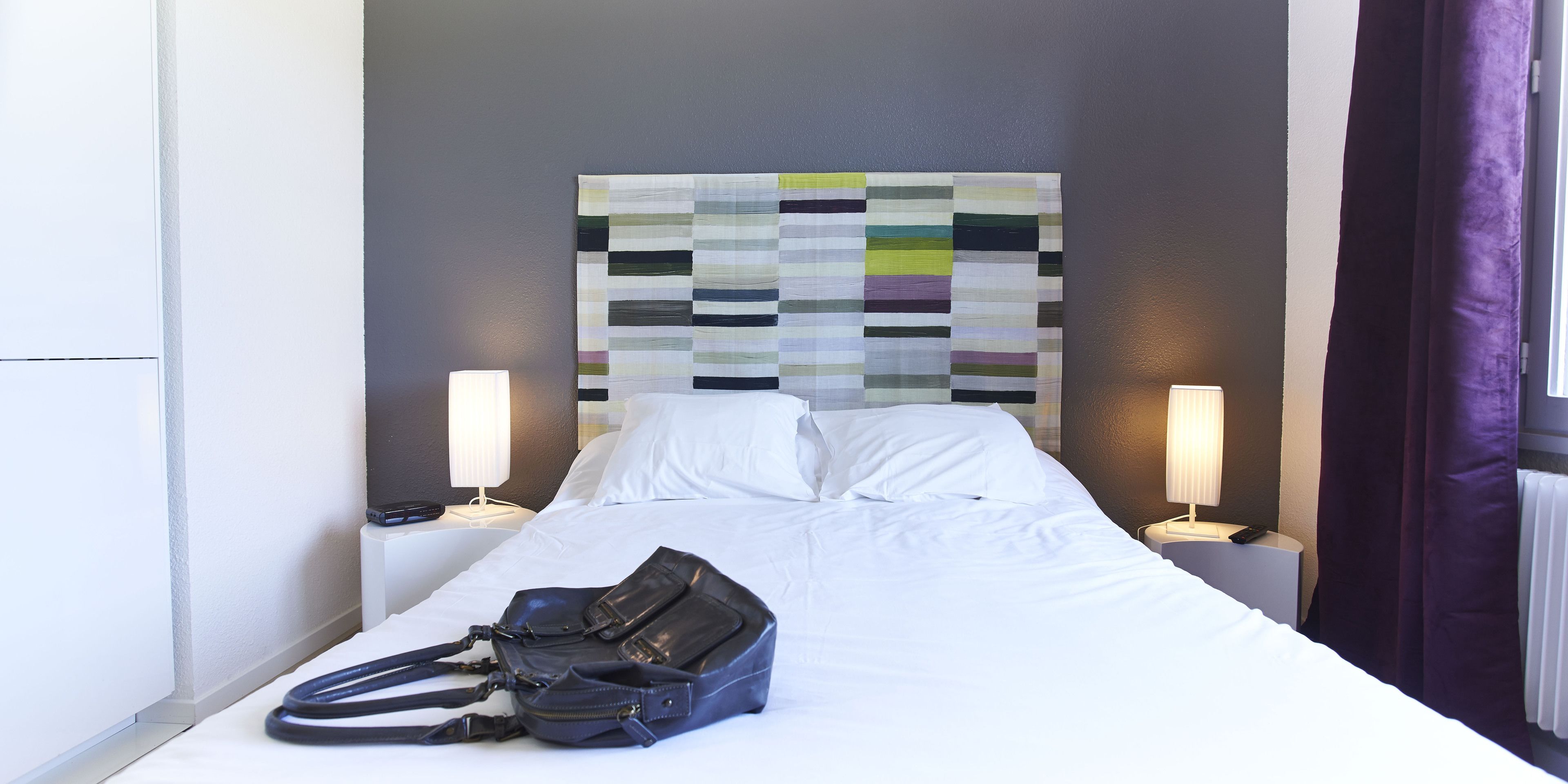 Een hotelkamer met een net opgemaakt bed, een hoofdbord met een geometrisch patroon, twee bedlampjes en een zwarte handtas op het bed.