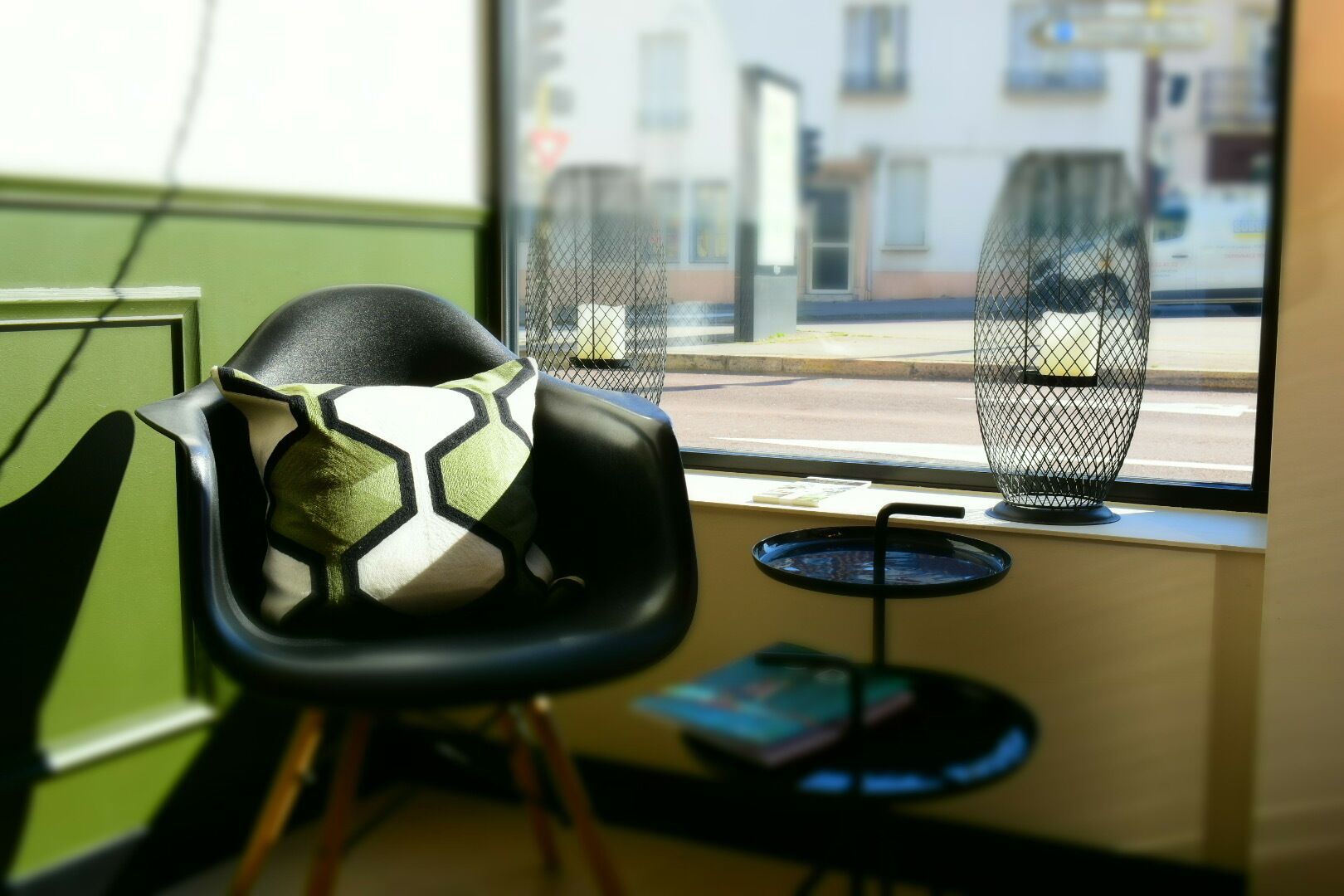 A chair with a geometric cushion, a small round table with a book, and a window with a view of buildings and a street.