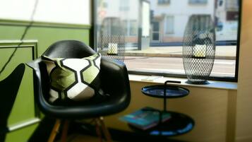 A chair with a geometric cushion, a small round table with a book, and a window with a view of buildings and a street.