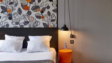 A hotel room with a bed, white pillows, a grey headboard, a wall with a floral pattern, an orange side table with a water bottle, and a black pendant light.