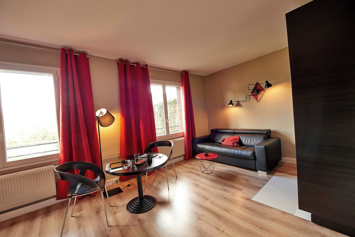 A modern living room with a black leather sofa, a dining table with chairs, a floor lamp, and red curtains.