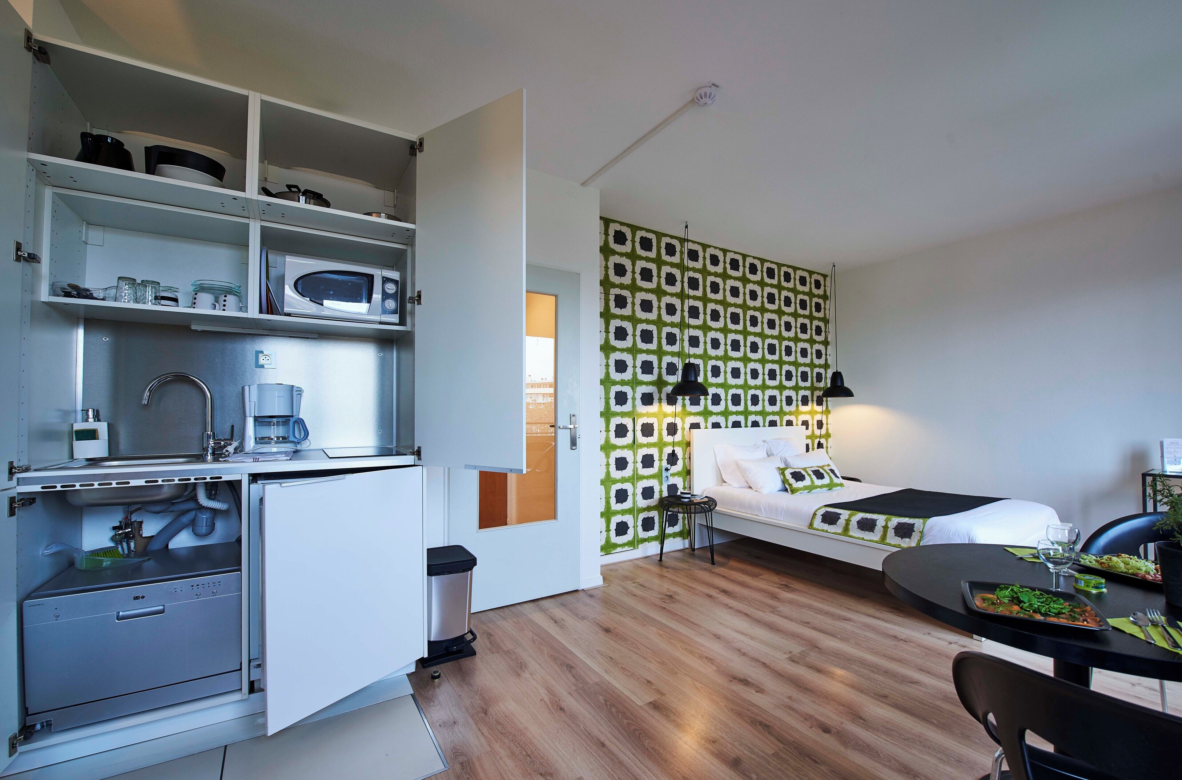 Een compacte hotelkamer met een kitchenette, een bed tegen een muur met patroon en een zithoek.